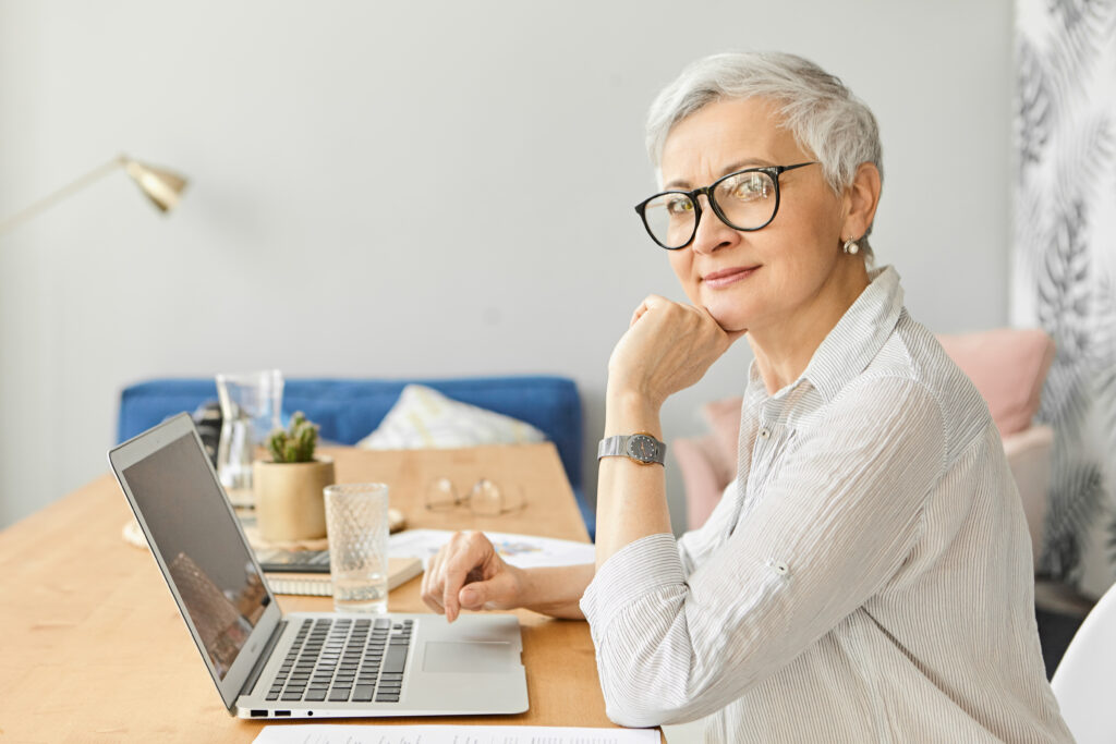 Modern electronic gadgets, occupation, age and maturity concept. Side view of attractive stylish middle aged self employed female in glasses sitting in front of open laptop, working at home office